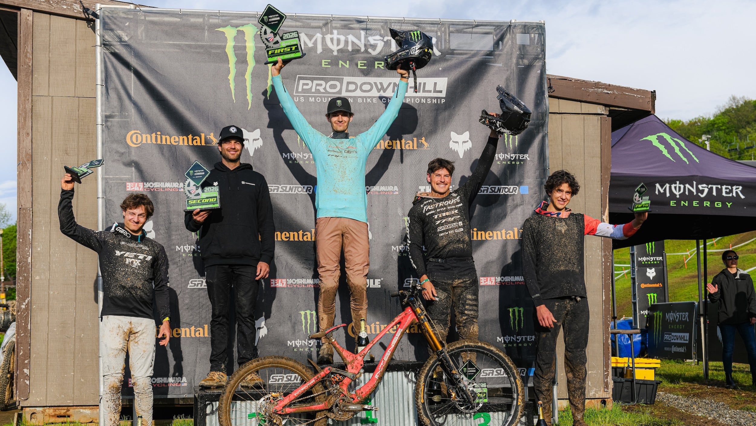 Biker on a podium with Monster Energy branding, holding trophies against a branded backdrop.