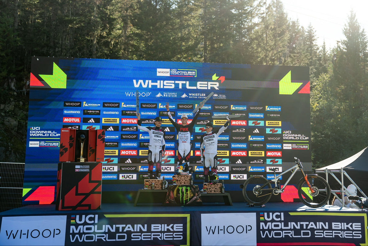 Three mountain bike riders stand on a podium at the UCI Mountain Bike World Series Whistler event, with a large branded backdrop behind them and a bicycle visible in the foreground.