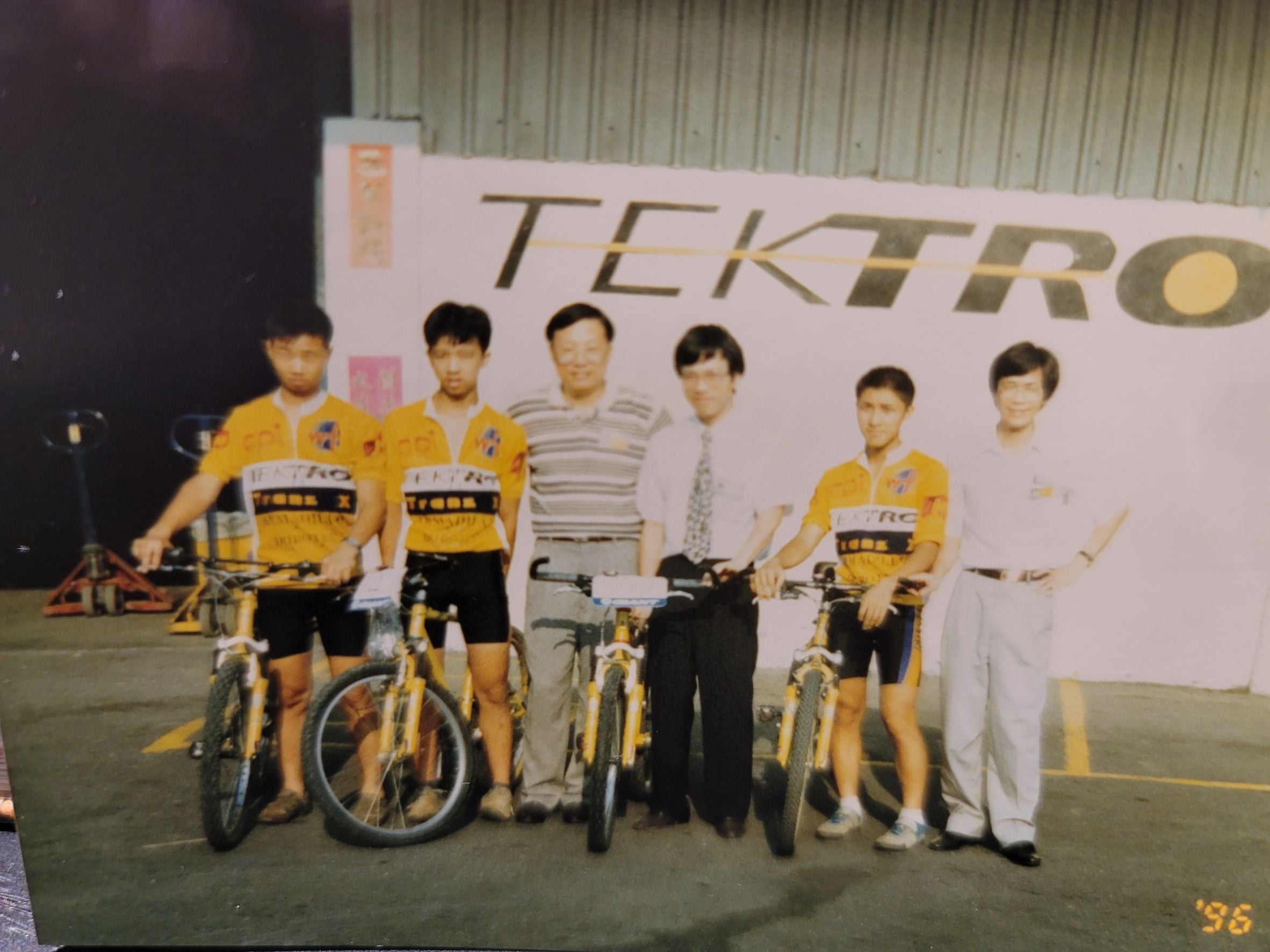 A vintage group photograph from the 1990s showing six men standing with bicycles in front of a large white TEKTO banner. Four of the men are wearing matching yellow and black cycling jerseys, while two men in the center wear casual shirts and trousers.