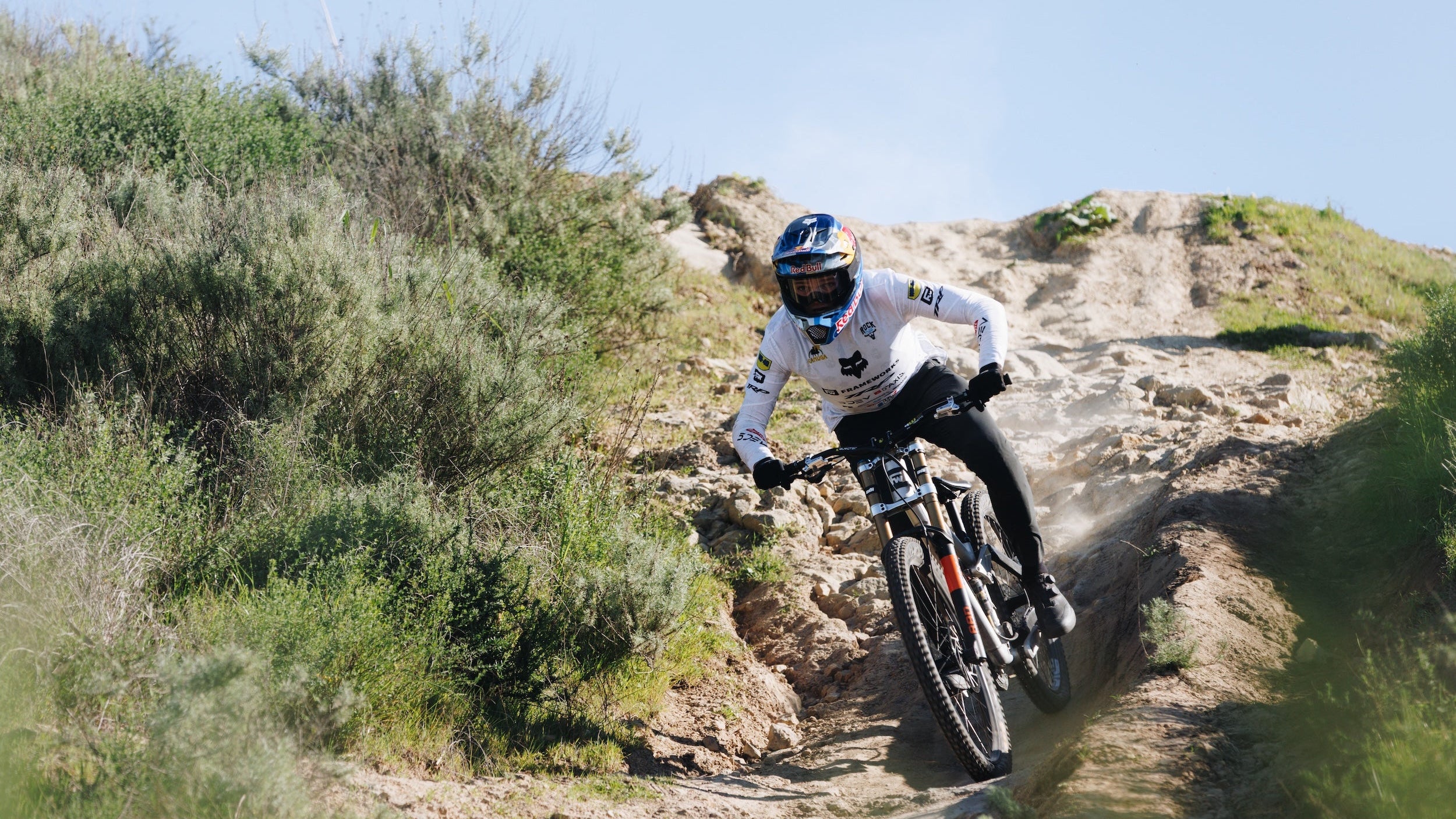 Person riding a mountain bike on a rocky trail with greenery and clear sky.