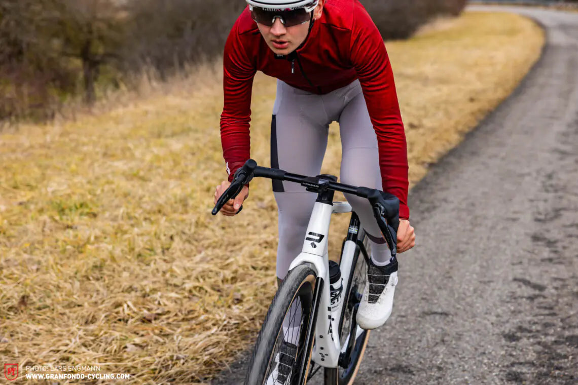 Cyclist in red jersey and gray leggings riding a white bicycle on a paved road.