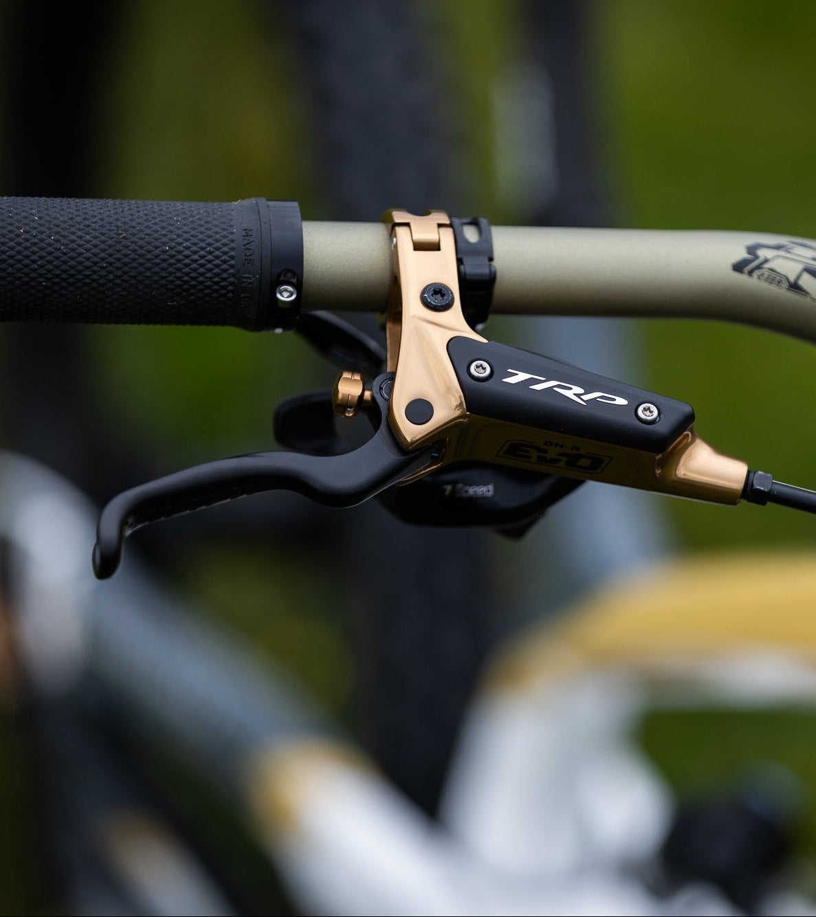 Close-up of a mountain bike handlebar with a black and gold TRP brake lever mounted on a tan handlebar, with a blurred outdoor background.