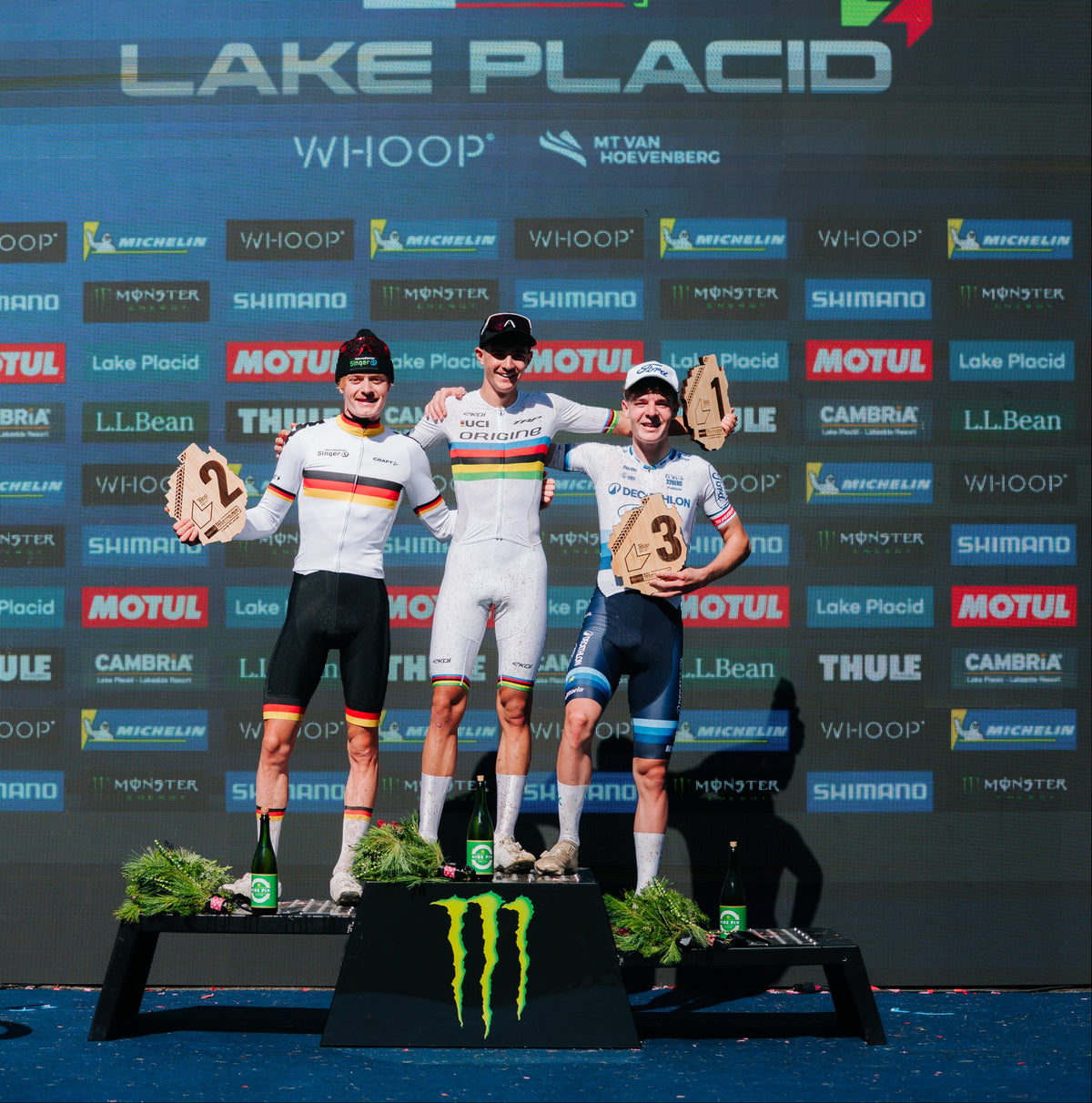 Three cyclists stand on a podium at Lake Placid, holding trophy cups. The backdrop features multiple sponsor logos including WhooP, Shimano, and Monster Energy.