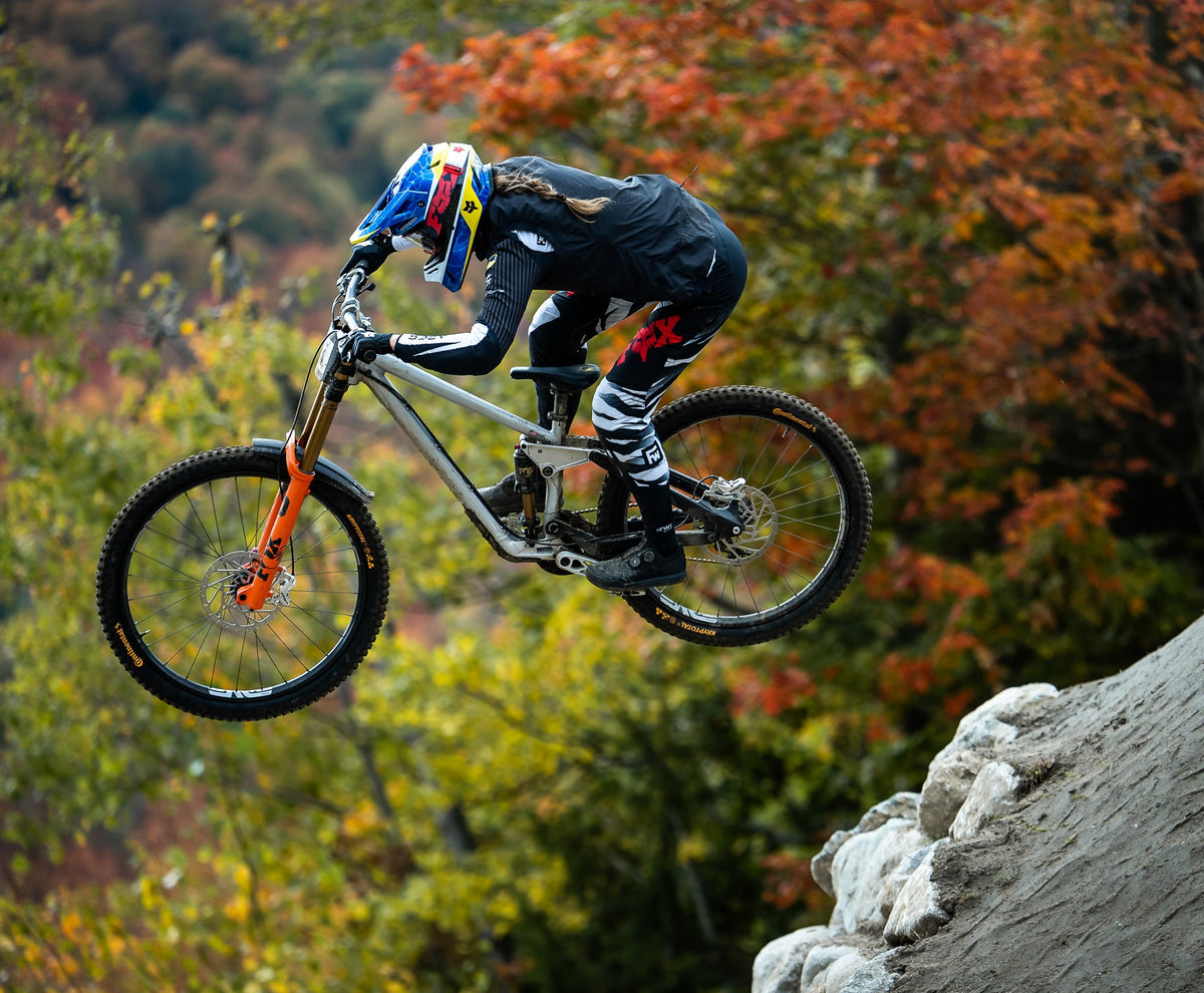 A full-suspension mountain bike with an orange front fork is airborne over a rocky drop-off, ridden by a motocross athlete in black gear and a blue, red, and yellow helmet, set against a blurred background of autumn foliage.