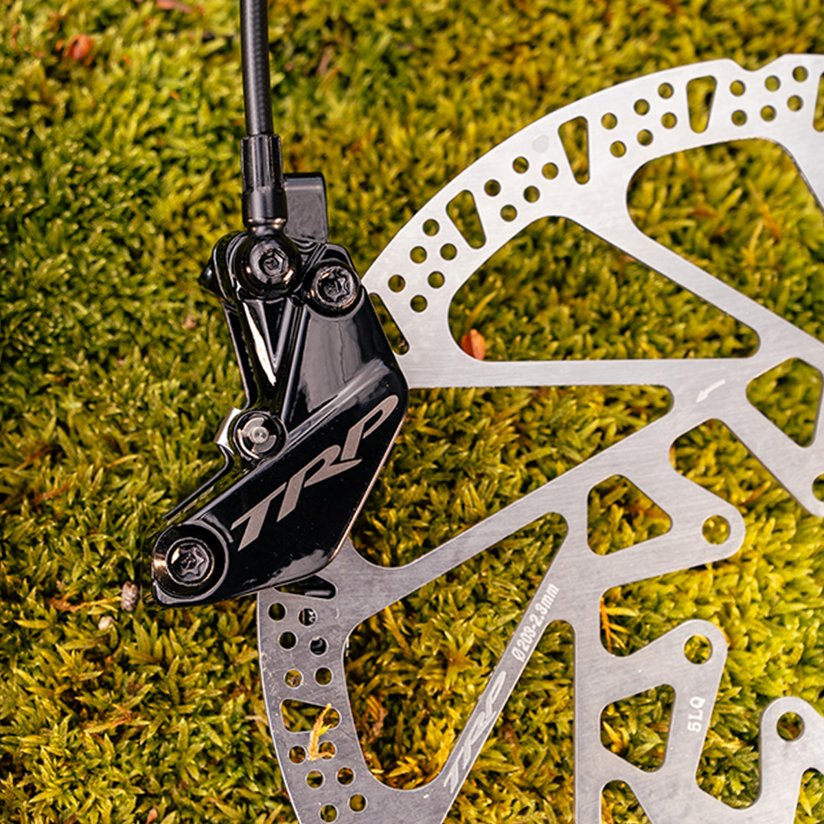 A close-up of a black TRP brake caliper mounted over a silver disc brake rotor, set against a background of green moss.