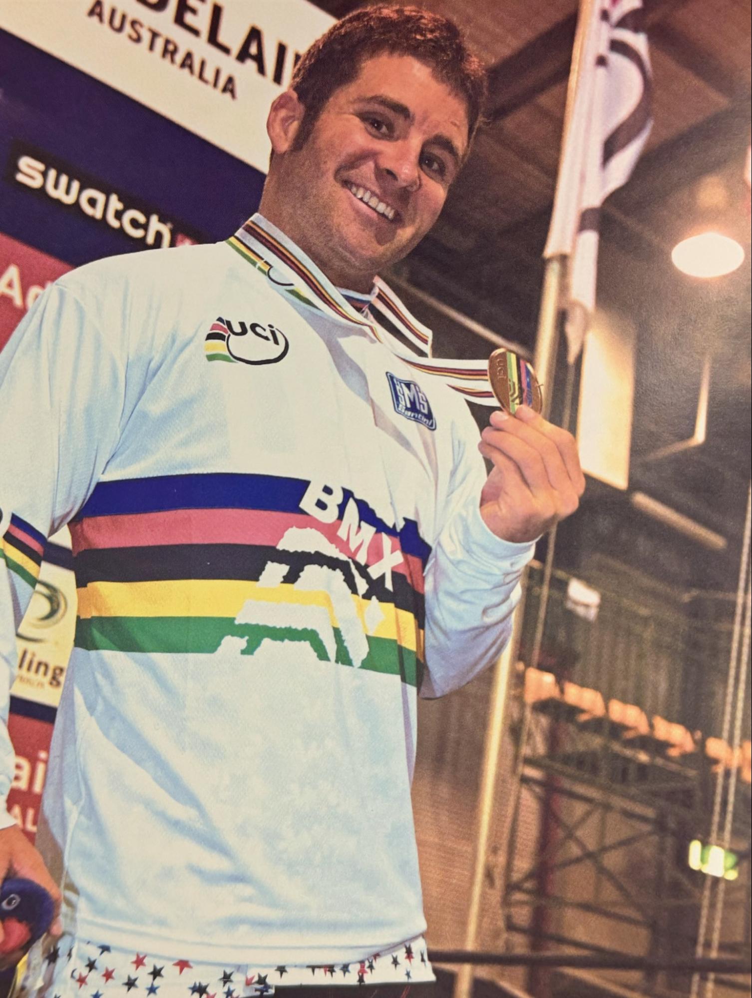 A smiling man in a white cycling jersey with a rainbow stripe across the chest and a medal hanging from his neck, standing indoors with a Swatch Australia sign in the background.