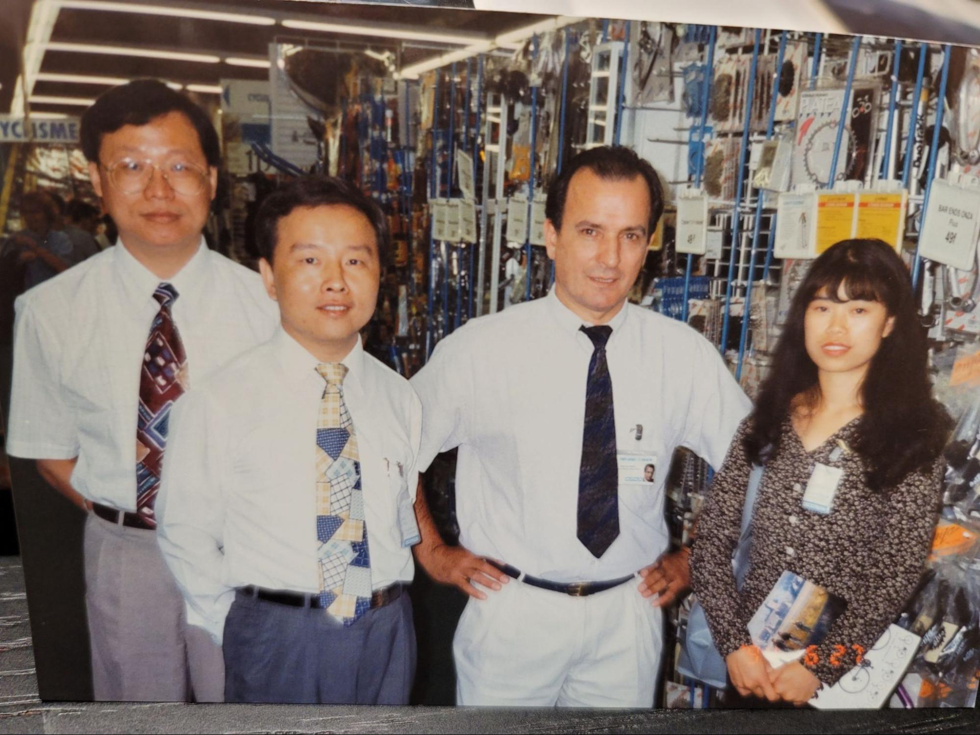 A group of four people standing together in front of a busy retail display filled with merchandise. The three men are wearing white shirts and ties, and the woman is wearing a patterned blouse.
