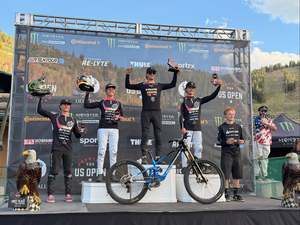 Four mountain bike riders stand on a podium at the US Open, holding trophies and helmets. A blue and black full-suspension bike is displayed in the center.